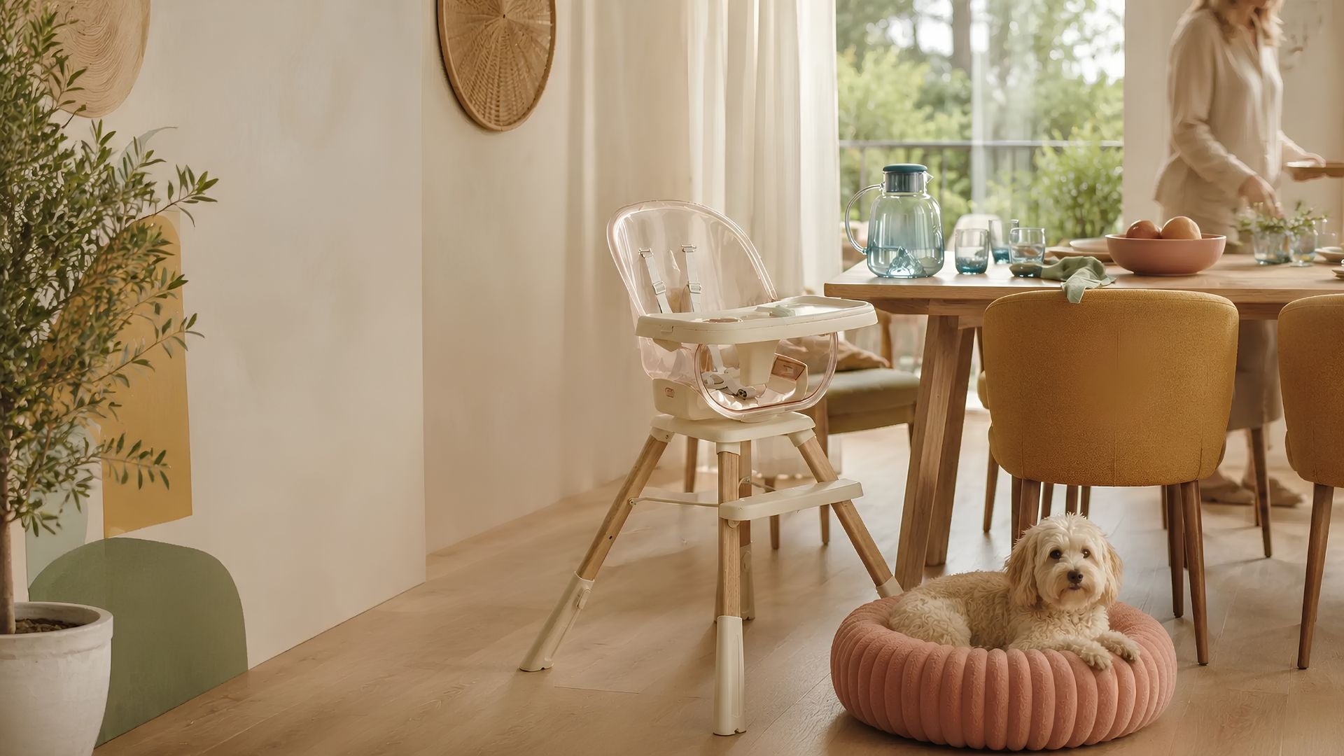 A baby high chair with a clear plastic seat and wooden legs stands in a bright, modern dining room next to a small dog resting in a pink pet bed. A woman is visible in the background near a wooden dining table.