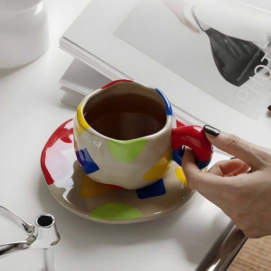 Bold abstract ceramic mug with colorful geometric shapes and saucer, held beside an open design magazine — by European House Hold
