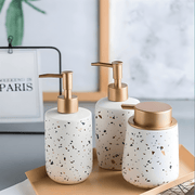 Trio of white matte ceramic soap dispensers with gold pumps in various geometric shapes on a bathroom tray.